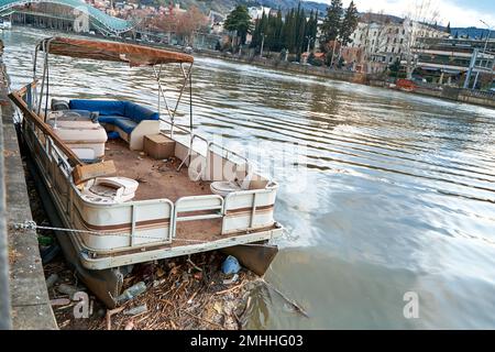 Ein verlassenes Freizeitboot legt sich am Ufer vor dem Hintergrund des Flusses Mtkvari und der Friedensbrücke in Tiflis nieder. Stockfoto