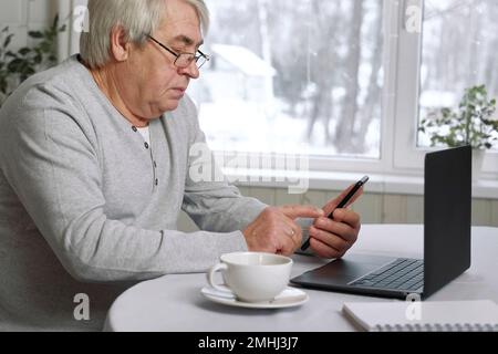 Ein gut aussehender, seriöser Seniorenmann, der eine Brille trägt und gleichzeitig mit seinem Laptop am Schreibtisch sitzt. Pensionierter Mann, der von zu Hause aus arbeitet Stockfoto