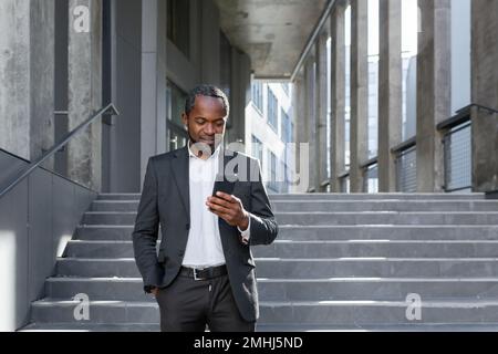 Ein erfolgreicher, reifer afroamerikanischer Mann in einem Business-Anzug geht aus einem Bürogebäude, benutzt ein Telefon, tippt eine Nachricht und sieht sich die Nachrichten online an, während er lächelt Stockfoto