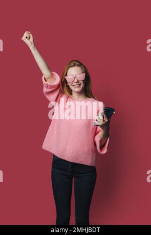 Fotoporträt einer aufgeregten, erstaunten Frau mit pinkfarbener Sonnenbrille, die das Halten des Telefons in einer Hand feiert, isoliert auf pastellfarbenem, karmesinfarbenem Hintergrund Stockfoto