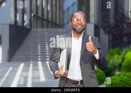 Porträt eines erfolgreichen afroamerikanischen Mannes vor dem Bürogebäude, Mann in Anzug und Brille, der steht und in die Kamera schaut, lächelt und Daumen nach oben gibt. Stockfoto
