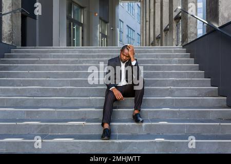 Ein aufgebrachter afroamerikanischer Mann, der vor dem Bürogebäude auf der Treppe sitzt, ein Geschäftsmann, der in Verzweiflung bankrott ist, ein reifer Boss, der in einem Geschäftsanzug im Freien deprimiert ist. Stockfoto