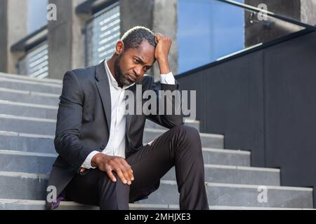 Ein aufgebrachter afroamerikanischer Mann, der vor dem Bürogebäude auf der Treppe sitzt, ein Geschäftsmann, der in Verzweiflung bankrott ist, ein reifer Boss, der in einem Geschäftsanzug im Freien deprimiert ist. Stockfoto