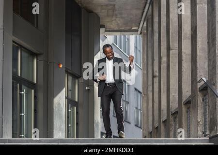 afroamerikanischer Boss feiert Sieg und Erfolg, Geschäftsmann tanzt auf der Treppe des modernen Bürogebäudes. Stockfoto