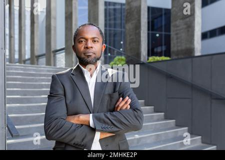 Ernsthaft denkender Geschäftsmann mit gekreuzten Armen, der in die Kamera schaut, Porträt eines afroamerikanischen Investorenbosses draußen im Büro, Mann im Business-Anzug. Stockfoto