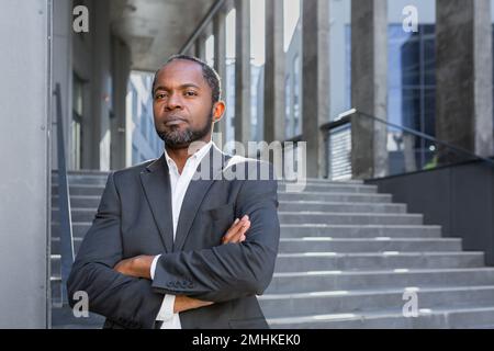 Ernsthaft denkender Geschäftsmann mit gekreuzten Armen, der in die Kamera schaut, Porträt eines afroamerikanischen Investorenbosses draußen im Büro, Mann im Business-Anzug. Stockfoto