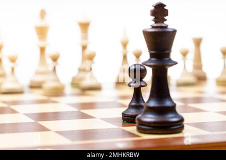 Schwarzer Bauer und Königin gegen weiße Schachfiguren im Hintergrund auf hölzernem Schachbrett (Fokus auf dem schwarzen Bauer) Stockfoto