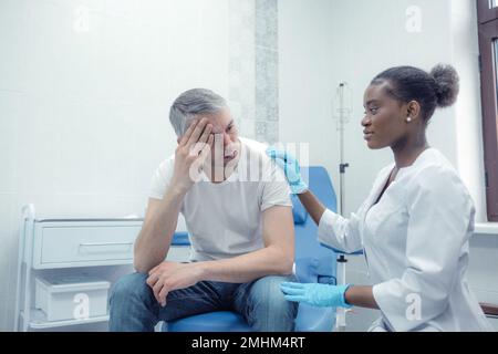 Ein unglücklicher erwachsener Mann, der unter Schmerzen, starken Kopfschmerzen, Stress und okulärer Migräne leidet, sitzt in einem Zimmer und bittet einen Arzt um eine Kontrolle und Hilfe bei der Behandlung Stockfoto