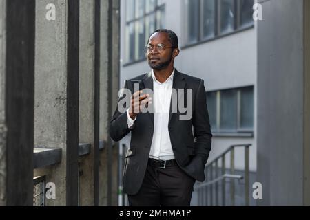 Seriöser und aufmerksamer Geschäftsmann, der Online-Nachrichten auf dem Telefon liest. afroamerikanischer Boss im Anzug außerhalb des Bürogebäudes, der die Konzentration des Smartphone-Bildschirms beobachtet, im Hintergrund in der Nähe eines Gebäudes läuft. Stockfoto