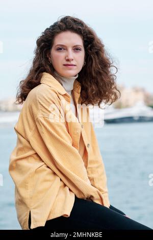 Junge Frau, die in die Kamera schaut, während sie sich draußen in der Nähe des Wassers posiert. Stockfoto