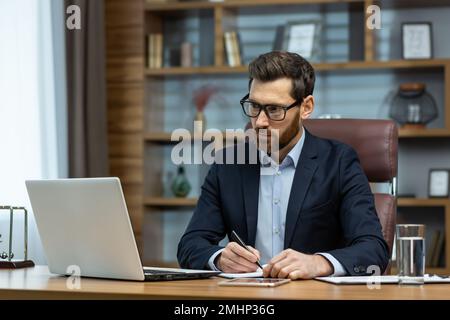 Erfolgreicher, erfahrener Geschäftsmann, der im Büro arbeitet, Mann im Geschäftsanzug, der am Arbeitsplatz mit Dokumenten und Laptop arbeitet, Chef hinter dem Papierkram, aufmerksam und fokussiert. Stockfoto