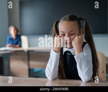 Die kleine Weiße langweilt sich beim Unterricht in der Schule. Das Schulmädchen sitzt an ihrem Schreibtisch und der Lehrer sitzt im Hintergrund. Stockfoto