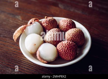 Tropical Fruit Lychee bekannt als Litchi chinensis Beeren auf dem Teller, dunkelbrauner Holzhintergrund, dramatisches Licht, Studioaufnahme. Stockfoto