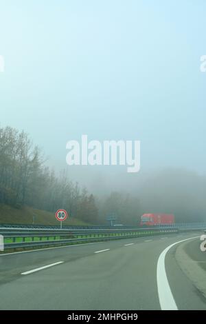 Auto fährt auf einer Autobahn, die mit starkem Nebel bedeckt ist. Roter Truck fährt von der gegenüberliegenden Spur. Transport. Schlechtes Wetter Stockfoto