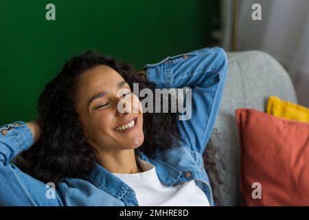 Junge hispanische Studentin, die sich nach dem Studium zu Hause auf dem Sofa entspannt. Sie legte ihre Hände hinter den Kopf, schloss die Augen und lächelte. Stockfoto