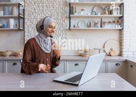 Junge araberin in einer Hidschab-Lehrerin, die zu Hause in der Küche sitzt, mit Laptop und Headset. Online-Unterricht, Gespräche per Videoanruf. Stockfoto
