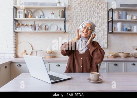 Junge arabische Studentin in Hijab, die zu Hause online mit einem Laptop studiert. Mit Kopfhörern am Tisch sitzen, Musik hören, ausruhen, Pause machen. Stockfoto