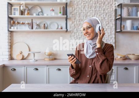 Porträt einer jungen muslimischen Frau in einem Hidschab, die zu Hause in der Küche sitzt, Kopfhörer trägt und ein Telefon in der Hand hält. Hört Musik, Podcast, Hörbuch, lernt Sprache. Stockfoto