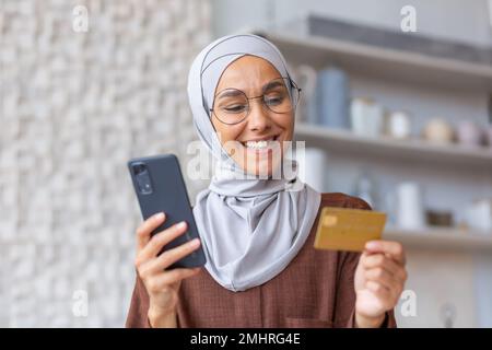 Nahaufnahme. Porträt einer jungen lächelnden arabischen Frau in Hijab zu Hause in der Küche, mit Telefon und Kreditkarte. Macht eine Bestellung, tätigt Online-Einkäufe, prüft das Konto, erhält ein Gehalt. Stockfoto