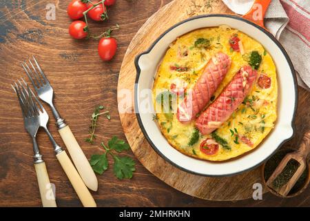 Omelette oder Frittata mit zwei Würstchen, geschmolzenem Käse, Kirschtomaten, Brokkoli und Petersilie auf roter Pfanne oder Eisenpfanne auf altem rustikalem Holzbacken Stockfoto