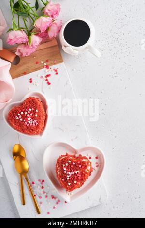 Zwei Kuchen Roter Samt in Form von Herzen auf weißem Teller, Rosenblumen und eine Tasse Kaffee auf rosafarbenem romantischem Hintergrund. Dessert-Idee für den Valentinstag, Stockfoto