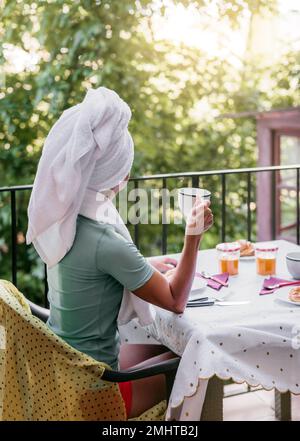 Eine weiße Frau mittleren Alters frühstückt nach einer Dusche auf der Terrasse ihres Hauses. Sie trägt ein Handtuch auf dem Kopf. Konzept des Wohlbefindens und Stockfoto