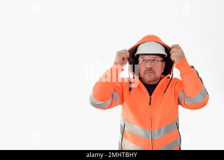 Ein Baumeister, der bei schlechtem Wetter eine Kapuze trägt. Ein Mann mit einem weißen Helm und einer orangefarbenen reflektierenden Jacke mit Kapuze. Weißer Hintergrund. Platz für Text Stockfoto