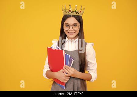 Schulmädchen in Schuluniform und Krone feiern den Sieg auf gelbem Hintergrund. Schulkind hält Bücher. Bildungsabschluss, Sieg und Erfolg Stockfoto