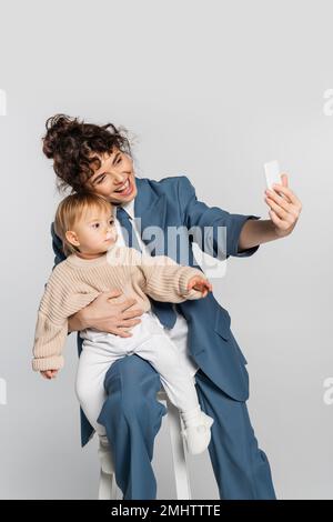 Glückliche Geschäftsfrau im blauen Anzug, die Selfie mit der kleinen Tochter auf dem Stuhl macht, isoliert auf grauem Stockbild Stockfoto