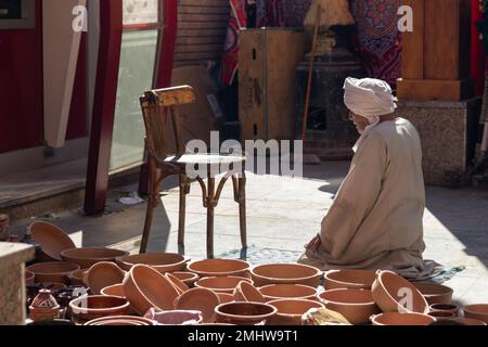 Ein unbekannter alter arabischer Mann mit traditioneller Kleidung, betend und auf den Boden blickend, während er handgemachte Tonschüsseln auf dem Straßenmarkt verkaufte Stockfoto