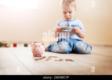 Süßer Junge, der drinnen auf dem Boden sitzt und Euro-Münzen in die Piggybank steckt. Stockfoto