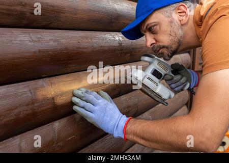 Reparaturarbeiter, der das Holzhaus mit einem Schleifgerät renoviert, bevor er den Fleck zum Wetterschutz aufträgt. Stockfoto