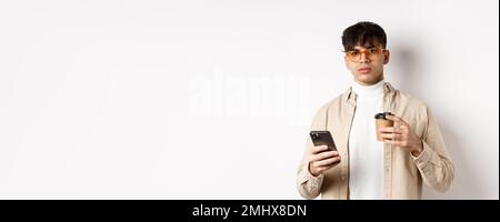 Echte Menschen. Stilvoller Hipster Kerl in Gläsern, die Tasse Kaffee vom Café zum Mitnehmen und Handy halten, auf weißem Hintergrund stehend Stockfoto