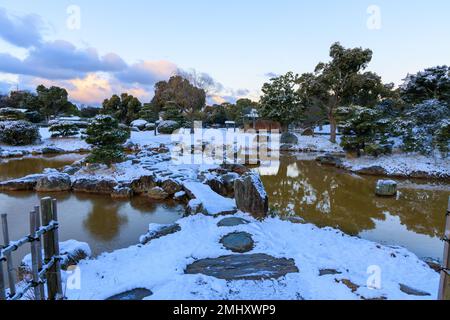 Morgengrauen im japanischen Garten mit Steinpfad und Brücke durch Schnee über den Teich Stockfoto