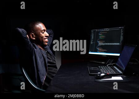 Entspannter männlicher Codierer, der sich zurücklehnt und in einem dunklen Raum den Code ansieht Stockfoto