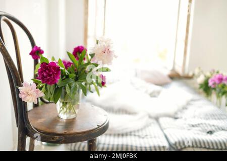 Nahaufnahme von Pfingstrosen in einer Vase auf einem alten Holzstuhl. Rustikale Einrichtung mit Matratzen, Kissen und Blumen in der Nähe eines offenen Fensters. Atmosphärisch m Stockfoto