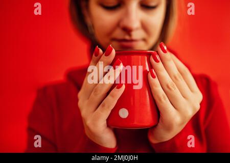 Ein blondes Mädchen in einem roten Pullover hält einen roten Becher in der Hand. Nahaufnahme einer wunderschönen, leuchtend roten Maniküre. Valentinstagskonzept. Kaffeepause. Rel Stockfoto