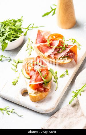 Offene Sandwiches mit Frischkäse, Prosciutto, Gurke und Rucola am weißen Tisch. Stockfoto