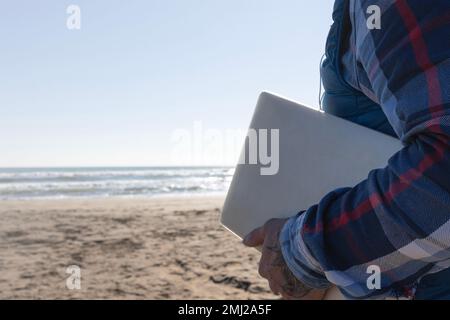 Die Hand eines Mannes hält ein Notebook am Strand, arbeitet im Freien, digitale Nomaden-Lifestyle-Konzepte Stockfoto