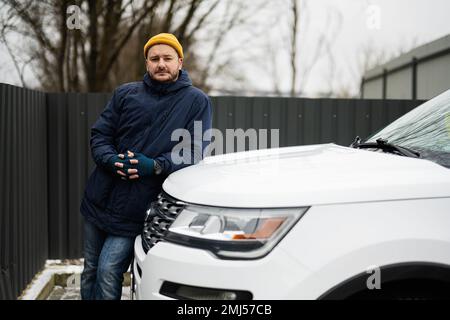 Man-Fahrerjacke und gelber Hut gegen die Motorhaube eines amerikanischen SUV-Wagens bei kaltem Wetter. Stockfoto