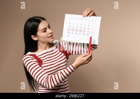 Junge Frau mit Kalender mit ausgeprägtem Menstruationstag auf beigefarbenem Hintergrund Stockfoto