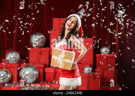 Schöne Frau im Weihnachtskostüm mit Geschenk unter Konfetti drinnen Stockfoto