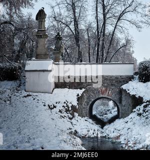 Schneebedeckte Steinbrücke mit zwei Statuen in Pardubice, Tschechien, zeigt historische Parkarchitektur, Winterlandschaft und eine ruhige Altstadt atmos Stockfoto