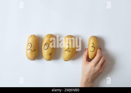 Vier rohe Kartoffeln mit der Nummer 2023 sind mit schwarzem Bleistift beschriftet, und die Hand einer Frau hält eine Kartoffel mit der Nummer drei auf weißem Hintergrund. Stockfoto