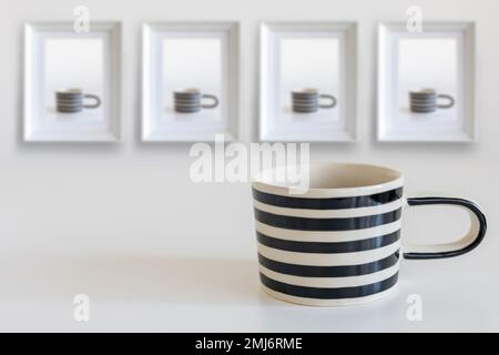 Tasse in schwarz-weißen Streifen, auf einem weißen Tisch. An der Wand hängen Fotos dieser Tasse in vier Rahmen. Vertikaler Rahmen. Platz für Text. Stockfoto