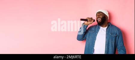 Schöner schwarzer Mann in Beanie und Jeanshemd, der Karaoke singt, Mikrofon hält und auf rosa Hintergrund steht Stockfoto