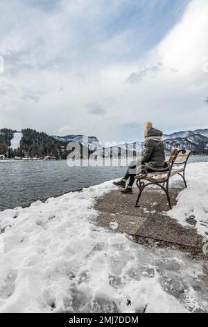 Frau sitzt auf einer Holzbank am Pier an einem bewölkten Wintertag vor dem Hintergrund des Bled Lake mit Schnee. Stockfoto