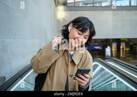 Porträt eines süßen koreanischen Mädchens im Trenchcoat, das die Rolltreppe hochfährt, das Handy hält und lächelt, pendelt, Videos ansieht oder Musik hört Stockfoto