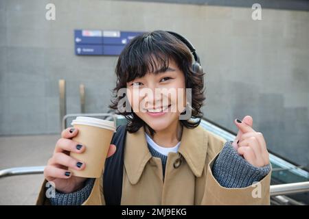 Porträt einer stilvollen asiatischen Frau in Kopfhörern, trinkt Kaffee zum Mitnehmen und lächelt, genießt Cappuccino auf dem Weg zur Arbeit, steht auf der Straße Stockfoto