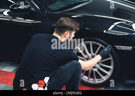 Rückansicht eines weißen Mannes mit schwarzen Handschuhen, mit Schwamm gebeugte, schwarze Autoreifen. Professionelle Autoverbesserung in einer Garage. Horizontaler Innenschuss. Hochwertiges Foto Stockfoto